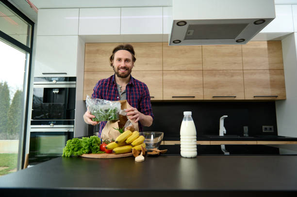 istock Handsome man smiles looking at camera, holding greens while unpacking eco paper bag with healthy food, standing at kitchen countertop in his spacious villa with large windows overlooking garden 1367887382