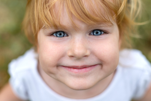 Happy smiling child face close up. Beautiful happy girl with blue eyes and blonde hair. Cute little kid smiling in a park, looking straight in camera close-up
