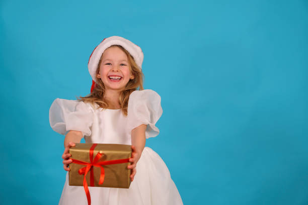 mignonne petite fille dans une robe de blom luxuriante pour des vacances. enfant amusant en chapeau de père noël tenir cadeau joyeux noël. belle fille sourit rire. boîte arc en or. bonne année !. place pour le texte. espace de copie. - blom photos et images de collection