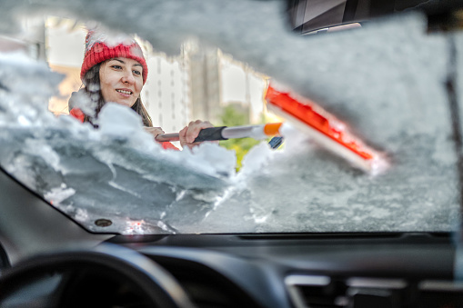 Millennial Woman in winter jacket scraping ice and snow from car windows