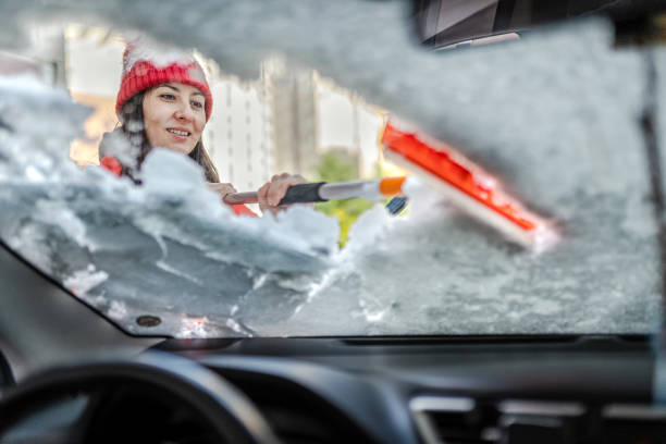 Millennial Woman in winter jacket scraping ice and snow from car windows stock photo