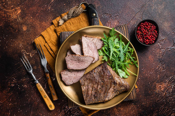 istock Roast Tri Tip or sirloin bottom beef steak on a plate with arugula. Dark background. Top view 1367896073