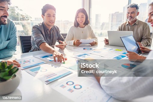 istock Multi racial diverse group of people working with Paperwork on a board room table at a business presentation or seminar. The documents have financial or marketing figures, graphs and charts on them. There are laptops and digital tablets on the table 1367899893
