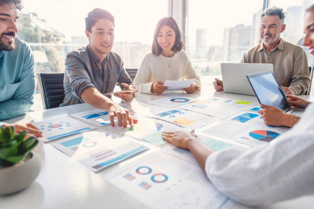 multi racial diverse group of people working with paperwork on a board room table at a business presentation or seminar. the documents have financial or marketing figures, graphs and charts on them. there are laptops and digital tablets on the table - analysera bildbanksfoton och bilder