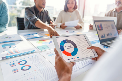 Group of people meeting with technology and paperwork. Close up of digital tablet with market analysis data on the screen. The documents have financial or marketing figures, graphs and charts on them. Multi ethnic group including Caucasian and African American