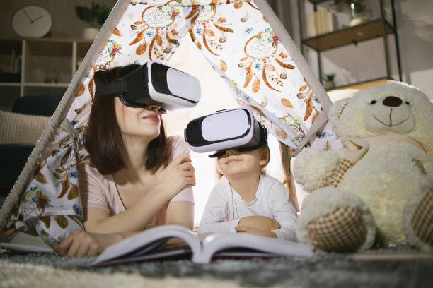 Happy mother and cute daughter lying in toy wigwam and using modern VR headset during evening time. Caucasian family of two using modern technologies for playing at home.