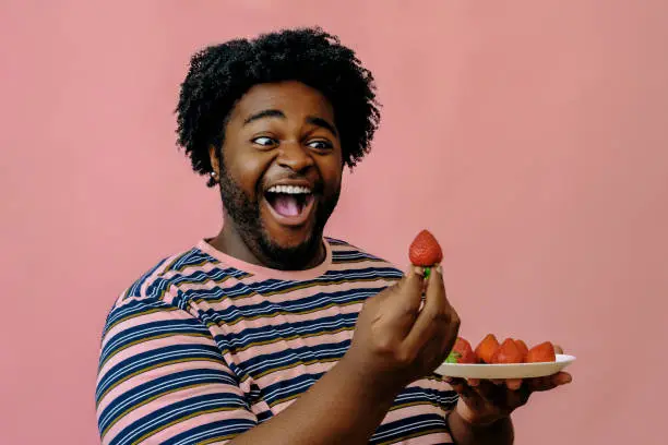 young happy african american man eating strawberries in the studio over pink background