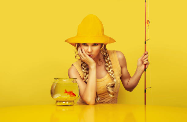 retrato de una joven pescadora aburrida, con la barbilla apoyada en la mano, vestida con un vestido amarillo con un patrón de pez, pescando un pez dorado en su pecera colocada sobre una mesa amarilla, sentada frente a un fondo amarillo. - fisherwoman fotografías e imágenes de stock