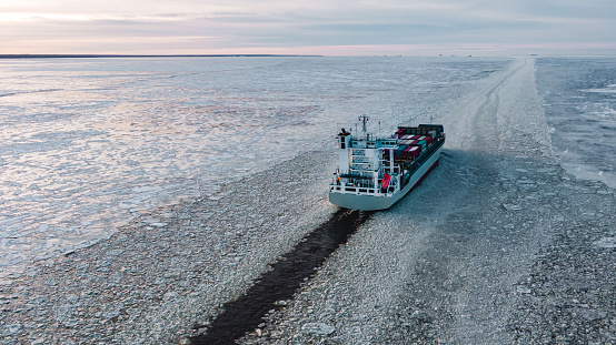 A large sea vessel with containers is moving in the ice on the sea along the fairway, aerial view