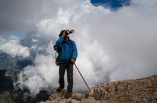 Mountain climber looks through binoculars enjoying spectacular view on top of the mountain above the clouds