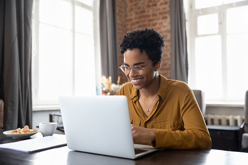 Happy young inspired African American woman in eyeglasses looking at computer screen, reading email with good news, enjoying studying or working distantly on online project at modern home office.