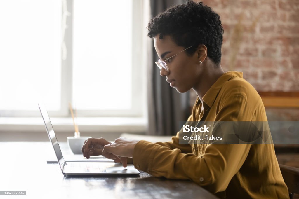 Focused young African American businesswoman working on computer. Side view focused young African American businesswoman in eyewear working on computer, sitting at table in modern loft office room, typing message, preparing electronic document or presentation. Using Computer Stock Photo
