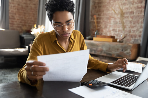 Focused young African American woman in eyeglasses looking through paper documents, managing business affairs, summarizing taxes, planning future investments, accounting alone at home office.