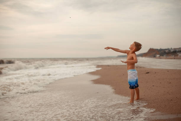A young perky tanned boy in shorts throws a small stone into the ocean on the shore during a storm on a summer day, space for text.