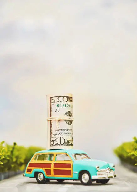 Photo of Station wagon car driving on a road with roll of money on roof. Savings and investment concept with copy space