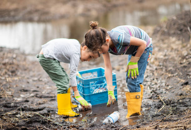 Cleaning Up for Earth Day Two children spend time on a mucky spring day as they clean up garbage and recyclables from the mud and puddles around them. They are wearing rubber boots and gloves as they work together to put items in a blue crate. earth day clean up stock pictures, royalty-free photos & images