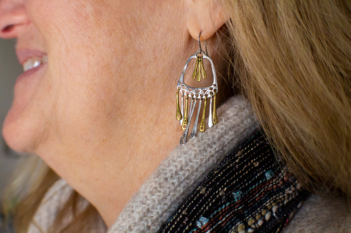 A close-up of a two-toned bronze and silver dangle earring on a woman's ear. The woman is smiling.  The model is wearing a multicolor scarf and a tan sweater.