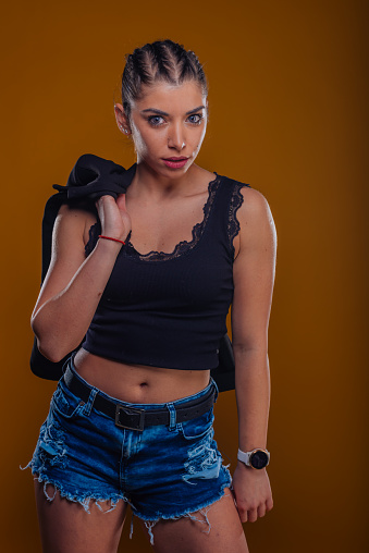 Young girl in denim shorts and black crop top posing on an orange light background.
