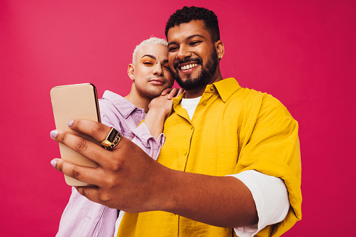 Cute gay couple taking a selfie together in a studio. Happy gay couple using a smartphone together while standing against a pink background. Young couple creating memories together.