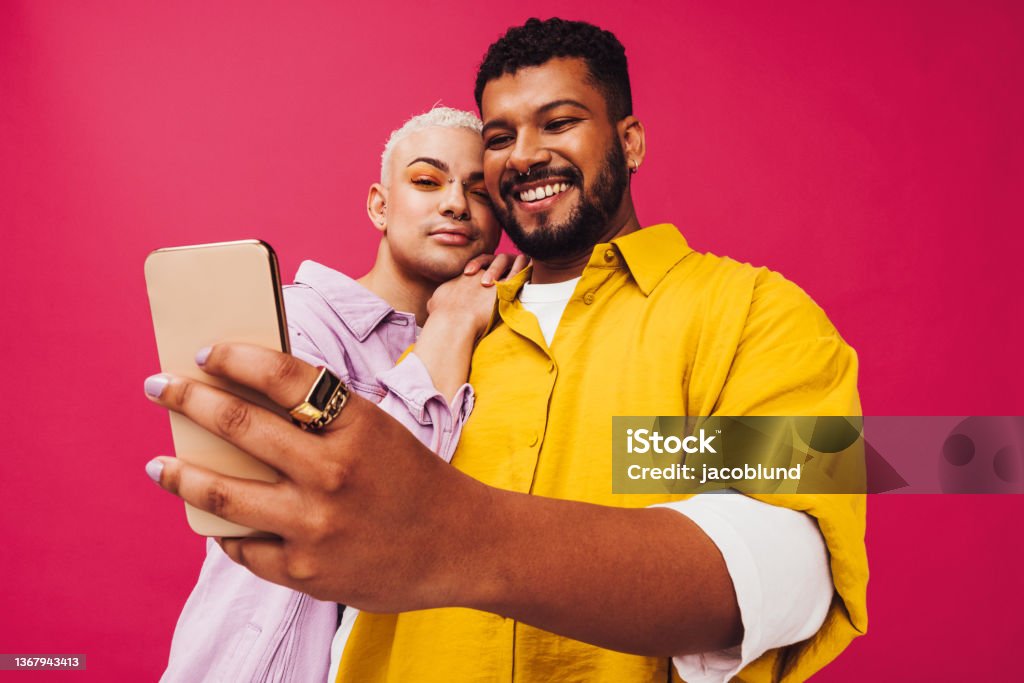 Gay couple taking a selfie together Cute gay couple taking a selfie together in a studio. Happy gay couple using a smartphone together while standing against a pink background. Young couple creating memories together. Generation Z Stock Photo