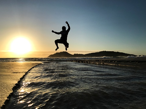 Young man jumping at dawn and backlit walking on the beach. Florianópolis city, Campeche beach, Santa Catarina