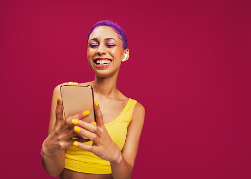 Vibrant playlists only. Happy young woman smiling cheerfully while holding a smartphone and wearing wireless earphones. Woman with purple hair enjoying streaming her favourite music in a studio.