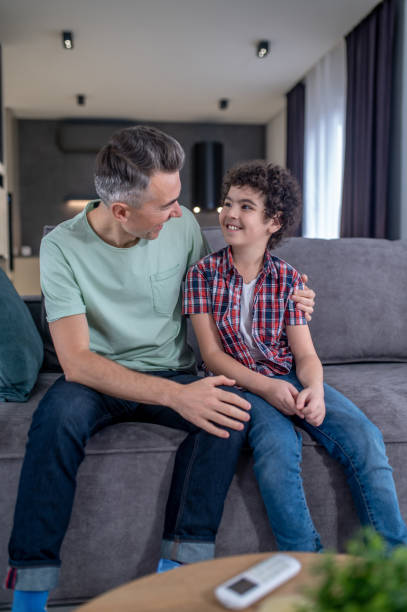 istock Man hugging boy sitting on sofa at home 1367953299