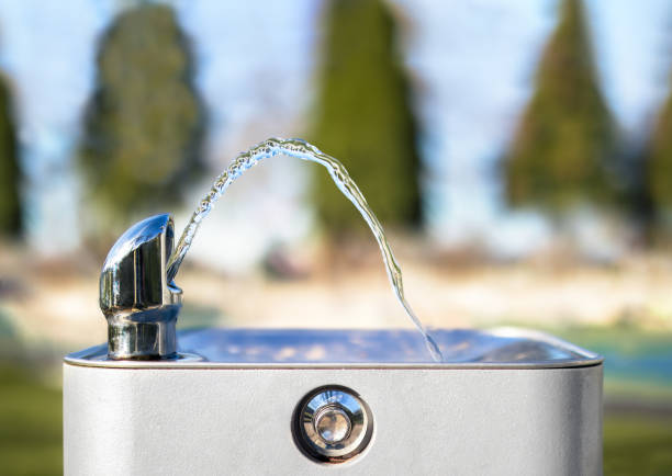 Drinking water fountain in park on a sunny day, no person. Close up of flowing water from the tab in an arch. Bright defocused park and tree background. Selective focus. drinking fountain stock pictures, royalty-free photos & images