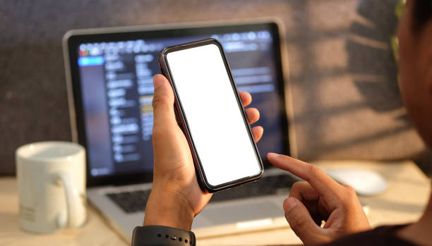istock Man sitting in front of computer laptop and holding smart phone with white screen. 1367956915