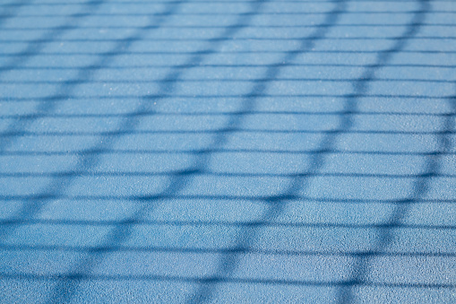 Defocused tennis net shadow on ground, outside early morning. Blue rubberized and granulated ground surface for shock absorption. Tennis sport backdrop texture.