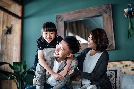 Playful young Asian family having fun playing at home while father giving daughter a piggyback ride in bed. Family life with love and happiness