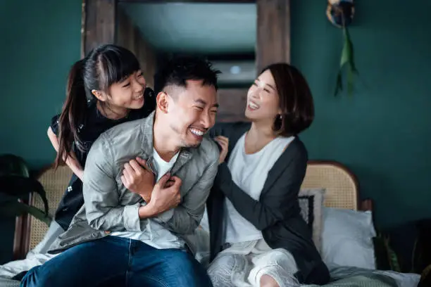 Photo of Playful young Asian family having fun playing at home while mother and daughter tickling father in bed. Family life with love and happiness