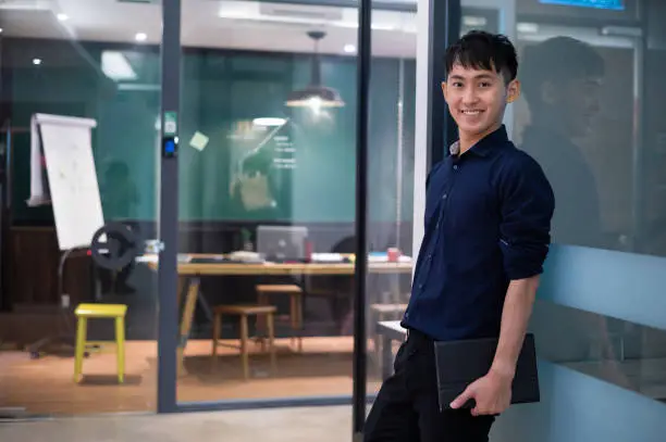 Portrait of young Asian entrepreneur businessman in modern office. Confidence male business executive with digital tablet in hands smiling and looking at camera.