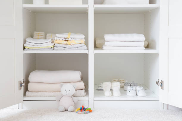 Different baby clothes, shoes and bedclothes on shelves in white opened wardrobe at nursery room. Front view. stock photo