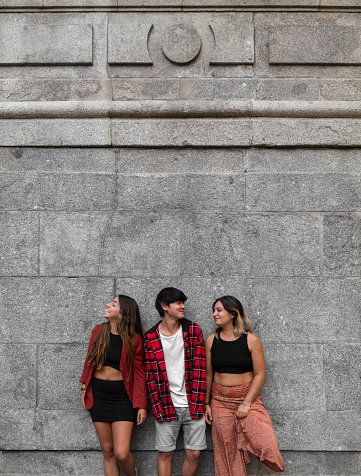 Portrait of two young girls and an Asian teenage boy laughing against stone wall. Madrid, Spain