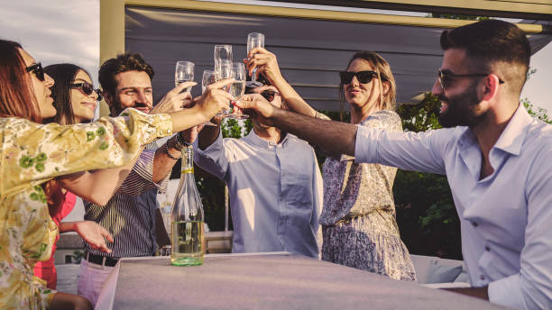 Laughing friends toasting wineglasses while sitting together. Cheerful group of friends celebrating outside a luxury hotel. Two happy couples enjoying a weekend getaway together. stock photo