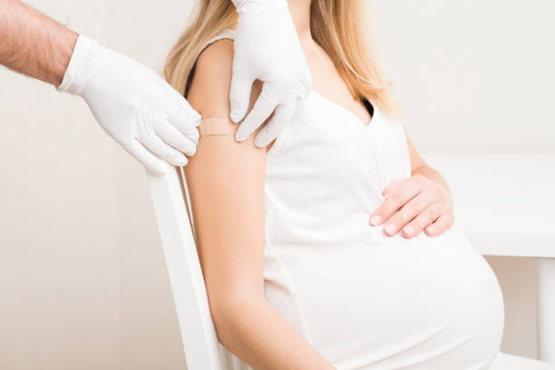 Pregnant young woman visiting physician at office. Doctor hands in rubber protective gloves putting adhesive bandage on shoulder after injection of vaccine. People vaccination and healthcare concept. stock photo