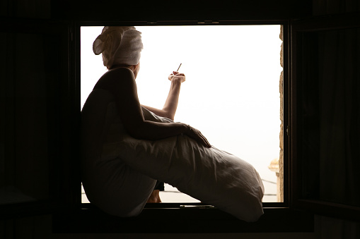 Woman with towel on her head, sitting in the window of a hotel room, smoking a cigarette