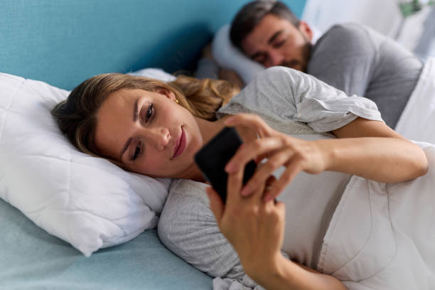 istock Young woman using mobile phone and checking messages in bed while her husband is asleep 1367973513