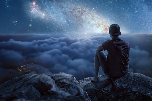 Man sitting on a rock above the clouds overlooking a beautiful night scenery with a starry sky.