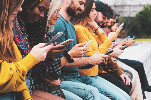 Closeup group of interracial gen z hipsters friends Sitting outdoors typing on  modern smartphone devices. 
Millenial people swiping on touch screens and using the internet concept
