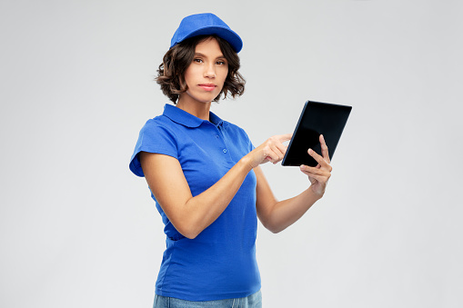 mail service, technology and shipment concept - delivery girl in blue uniform with tablet pc computer over grey background