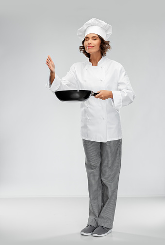cooking, culinary and people concept - happy smiling female chef in toque with frying pan smelling food flavor over grey background