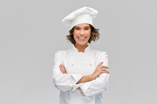 cooking, culinary and people concept - happy smiling female chef in toque with crossed arms over grey background