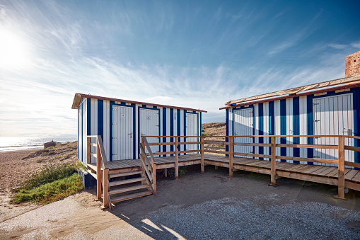 A small cabin with toilets at the coast. The cabin is blue and white colored and has a little red roof. Behind it you can see the beach and the sea.