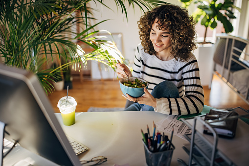 Curly young woman sitting in office at home, she is eating a salad at the desk.