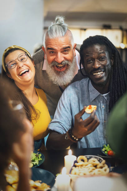 multi-ethnic group of friends smiling looking at camera while they're dining together sitting at home stock photo