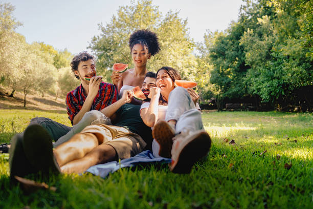 gruppe glücklicher junger leute, die spaß daran haben, wassermelone beim picknick zu essen und auf dem gras zu sitzen. lebendige warme farben gefiltertes bild. - family african ethnicity black african descent stock-fotos und bilder