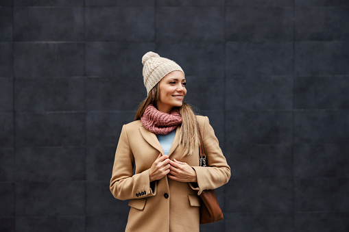 Feminine fashion and woman style. A stylish happy young woman in a coat, scarf, and beanie standing outdoors and buckling up the button.