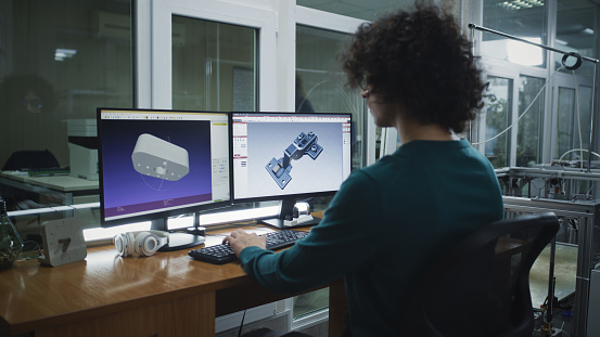 Young man in casual clothes with curly hair creating 3D model on computer while sitting at desk in modern design studio
